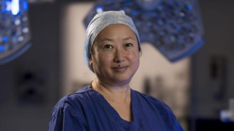 Evelyn Ong is standing in an operating theatre wearing dark blue surgical scrubs and her hair is encased in a wrap.