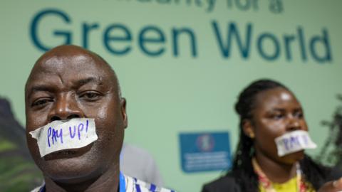 Two people wear tape over their mouths that reads "Pay up" in front of a sign that reads "Solidarity for a Green World" at COP29 in Baku, Azerbaijan