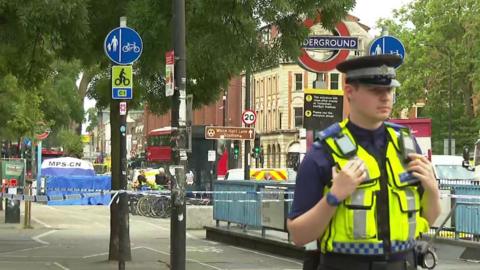 A police officer stands by a cordon at the scene, with Tube station roundels and a blue and white forensic tent in the distance