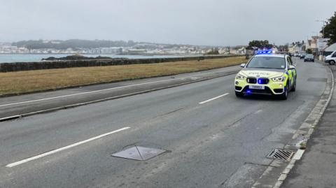 A police car with flashing blue lights drives along a coastal road with a town in the far distance across the water.