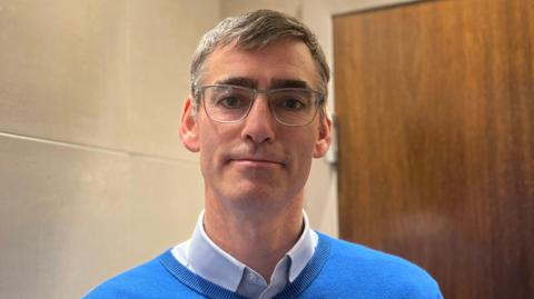 Monty Tadier has short greying hair and is wearing glasses, and a blue jumper with a white shirt underneath.