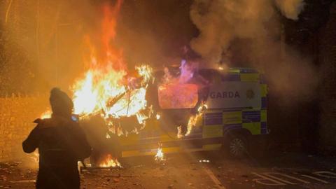 A police van is on fire. There is a person standing in front of it, they have a hat on. The front half of the van is almost entirely burnt. The background is a blur of flames and smoke. There is a stone wall behind the van. 