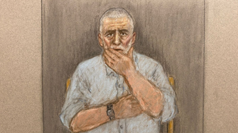 A court sketch of Peter Sullivan. He has grey hair, a grey beard, and is wearing a grey shirt. He has one arm folded across his chest and the other is to his face. 