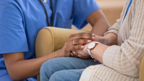 A woman's hands holding another person's hands. The woman is wearing a blue nurse's uniform and the other person is sat on a chair. 