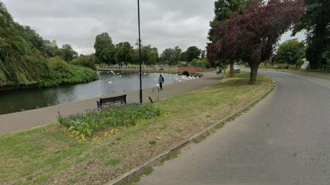 The River Nene as it curves round to the left, with a promenade pavement, strip of lawn, and road all following the same curve. There are swans on the river in the distance and you can see a small brick bridge that runs alongside the river. There are large trees either side of the river and a playground in the distance.