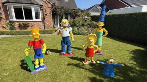Five statues depicting Bart, Homer, Marge, Maggie and Lisa from the TV show The Simpsons stand on a green lawn. A brick house is in the background.