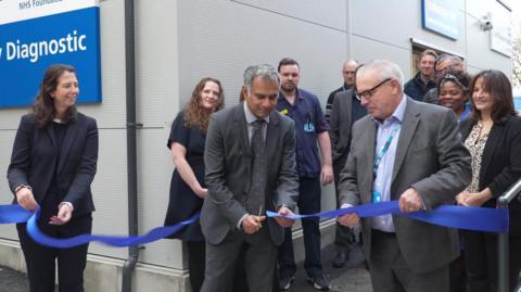 Two men wearing suits stand close to each other in front of a building. The man on the right is holding a blue tape, which is being cut in a ceremonial fashion by the man on the left. On the far left is a woman also holding the tape and there are a number of people standing watching in the background.