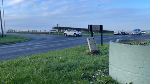 A road junction which has a stone sign like a fire hydrant which has been partially knocked over. There is a green planter in sight and several cars.