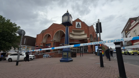 Police tape stretched between lamp-posts and other street furniture with an ornate blue clock in the middle of a town square and late 20th Century brick shopping centre behind.