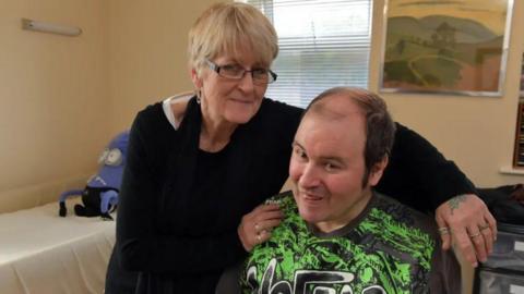A woman with short blonde hair and black glasses, wearing a black jumer is seen with her arms around a man who is sitting down.  The man has brown hair, thinnin gon top and wearing a green and black sports top.