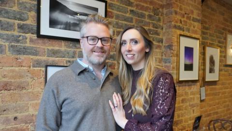 A woman and a man stood next to each other in a exposed red-brick room.