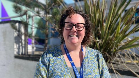 A woman with curly brown hair and black glasses. She is wearing a blue and green top with a light green vest. She is wearing a blue lanyard which says "Guernsey Police" on. Behind her is a white wall and green bush. 