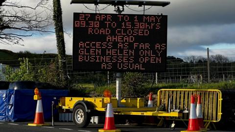 A large portable matrix sign on a yellow trailer with the details of the closure on it. There are traffic cones and crash barriers around it.