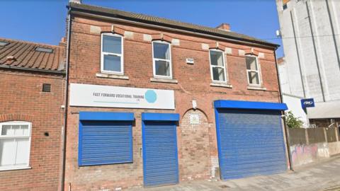 The exterior of the Fast Forward Vocational Training offices. It is a red brick building with three sets of blue shutters. There is a white sign above two of the shutters, which seem to cover a window and a door, that reads "Fast Forward Vocational Training" in capital letters. There is a grey path in front of the building. The sky is blue.