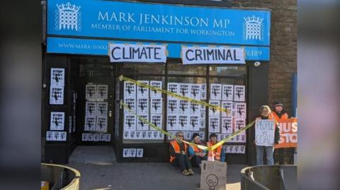 Just Stop Oil protesters sitting outside Mark Jenkinson's office. They are wearing hi-vis vests. The windows of the office are covered in posters which read "Stop Tory Coal" and a larger banner saying "Climate Criminal" is attached to the area above the windows.