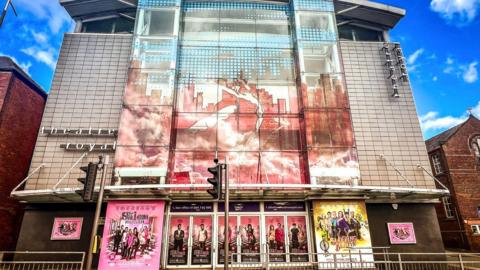 The front of St Helens Theatre Royal, which is adorned with posters for upcoming events. It is a tall white-tiled building with glass frontage.