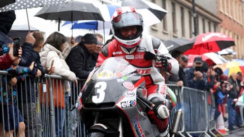 A motorbike rider, dressed in red and white protective gear, is on a racing bike with the number 3 on the front. He has a helmet on but you can tell he is smiling at the camera. There are rows of people behind him in the street. They are separated by grey metal dividers. A number of the spectators have umbrellas up.