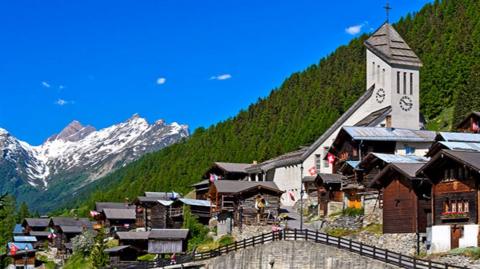 A general view of a row of buildings in Blatten, on an elevated road overlooking a stream. There are forest-covered mountains behind. The sky is bright blue and there are snow-topped mountains in the background.