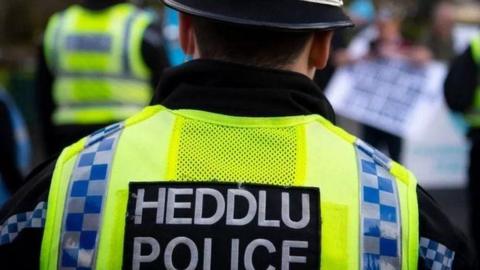 A police officer wearing yellow, blue and black uniform and a hat, with their back to the camera. Their vest reads 'Heddlu, Police'.