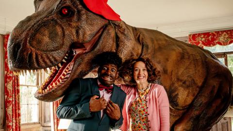 two people, a man and a woman, standing in front of a t rex wearing a red hat