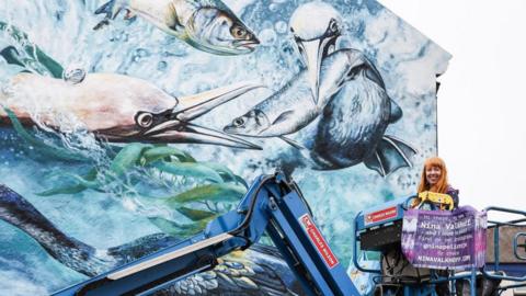 Giant artwork on the side of a building depicting an underwater scene with two seabirds and two fish. To the right is the artist standing in the cage of a cherry picker. She has long reddish hair and is smiling.
