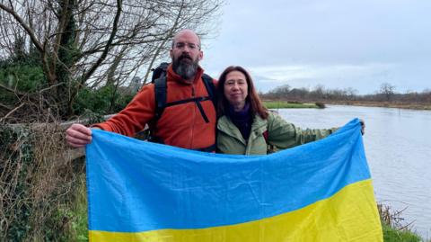 A man with a beard in a red coat stands next to a woman with a green coat. The pair are holding up a blue and yellow Ukraine flag. 