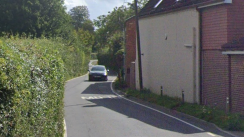 Black car driving down a narrow road. The road has a SLOW road marking. The road is surrounded by bushes. The vehicle will be driving by a building on the right hand side of the picture.