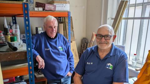 Two men in their seventies in a small box room filled with DIY tools, shelves and wood. Both are wearing blue tops with a 'WM' logo on and their Christian names printed across the right side of their chests. 