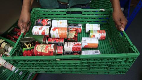 Close up of a green plastic basket with tins of tomatoes in it being held by a foodbank volunteer