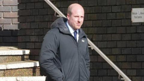 A bald man in a navy suit with a black coat on top walking down stairs.