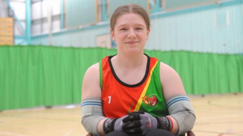 Sarah Burrows, captain of Penrith Pumas wheelchair rugby team. She has light brown hair tied back and blue eyes. She is inside a sports hall and is wearing a red, yellow and green vest and protective sport equipment including gloves and elbow pads.