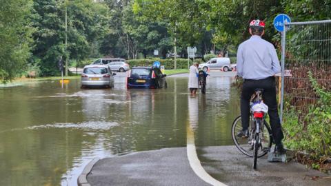 A flooded road with two cars stuck in the middle of it. A man on a bike is surveying the scene on a pavement above the water. Another woman is trying to push her bike through the flooded road