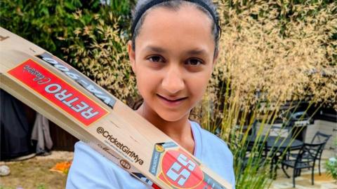 A young girl with dark hair tied up in a ponytail and wearing a white T-shirt. She's resting a cricket bat on her shoulder and looking straight at the camera. She appears to be in a residential garden, with plants and a table and chairs behind her.