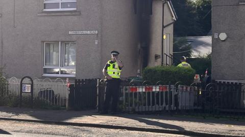 Police and utility workers stand outside a residential corner property on Cumbernauld Road. A marked service van with its rear doors open is parked at the roadside, while barriers surround the entrance to the property. The scene is photographed in daylight with neighbouring houses and trees visible in the background