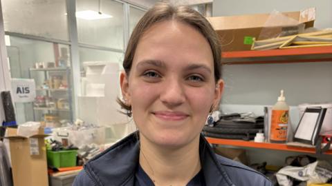 Millie Brown, a young lady with brown hair in a ponytail, smiles from inside an engineering lab at her university. There are busy-looking shelves and piles of equipment, boxes and other items behind her.