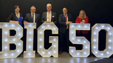 Three men in suits and two women stand behind some illuminated giant letters spelling out "Big 50".