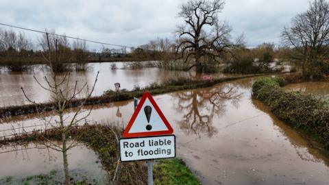 Flooded fields and impassable roads with tree-trunks submerged in water and signage explaining the road is liable to flooding. 