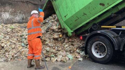 A man in orange overalls stands in front of a pile of rubbish at a tip. On his right a lorry tips waste into the pile