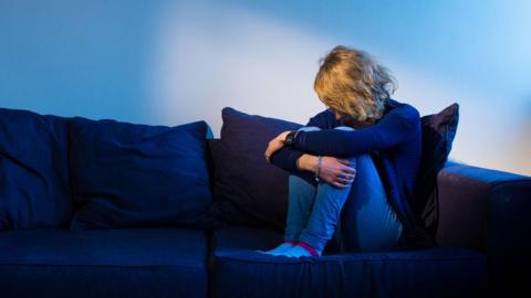 A woman with blonde wavy hair sitting on a navy blue sofa hugging her knees to her chest with her head bowed forward