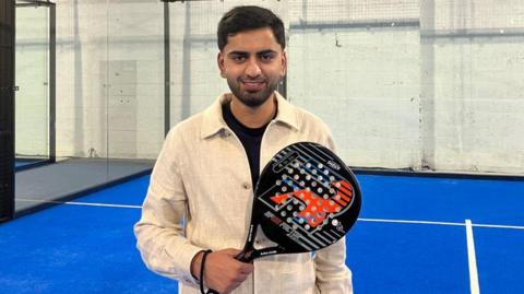 A man in a cream overshirt holding a black padel racket is smiling on an indoor padel court