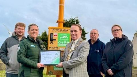 A group of five people in a line standing in front of the yellow defibrillator box. Megan Stephenson is in the middle left, in a green NWAS uniform and holding a certificate alongside the mayor who is middle-right. He is wearing a checked blazer and has the ceremonial chain around his neck. 