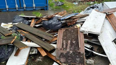 A pile of rubbish sits on the ground in front of a large blue shipping container. Rubbish includes planks of wood and black bin bags