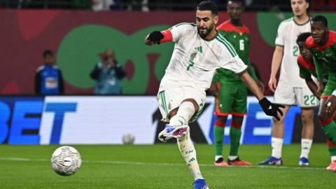 Footballer Riyad Mahrez, wearing a white jersey with green stripes and the number seven on it while playing for the Algeria national football team, kicks the ball while taking a penalty against Burkina Faso at the Afcon 2025 tournament, with opposition players in a green kit and red trim visible in the background