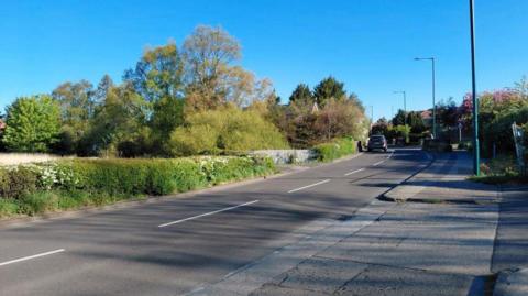 Hutton Lane - a road in a suburban area, with farmland along one side and homes on the other. A hedge marks the edge of the farmland, behind which stands tall grass and trees.It is a clear and sunny day.