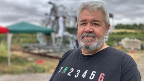 Fraser Drummond. He has short grey hair and a beard. He is stood in front of the sculpture, which is out of focus in the background. Fraser is looking at the camera and smiling.