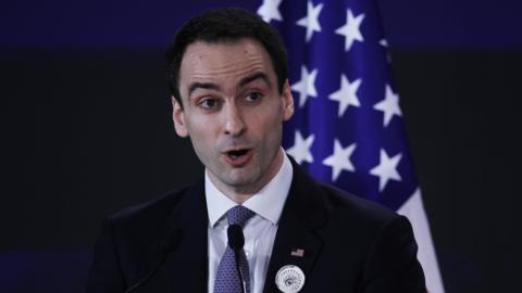Michael Kratsios, a White House director and advisor on technology, speaking into a microphone at a podium, wearing a black suit jacket, white dress shirt and blue patterned neck tie. An American flag is positioned upright behind him. 