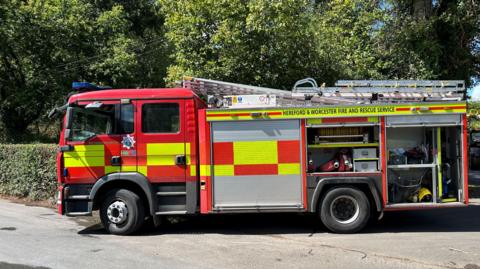 A fire engine in a countryside lane. The engine is mainly red with yellow rectangles on the side. A ladder is on the roof.