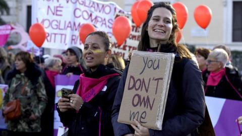 Two people stand during a protest, one smiles and the other looks away from camera holding a placard that reads 'you don't own me', with other demonstrators in the background holding orange balloons, in Rome on 22 November.
