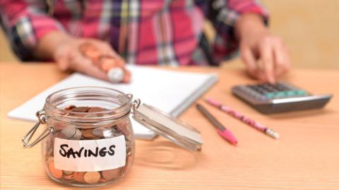 Jar of coins with sticker saying "savings" in the foreground, with a hand, coins, calculator, pen, pencil and paperwork behind.