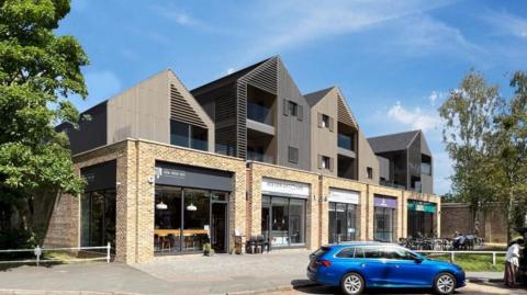 An architect's image of proposed flats. At street level there are shop fronts and above them are flats with pointed rooftops. There is a car parked in front of the buildings and a grassed area to one side